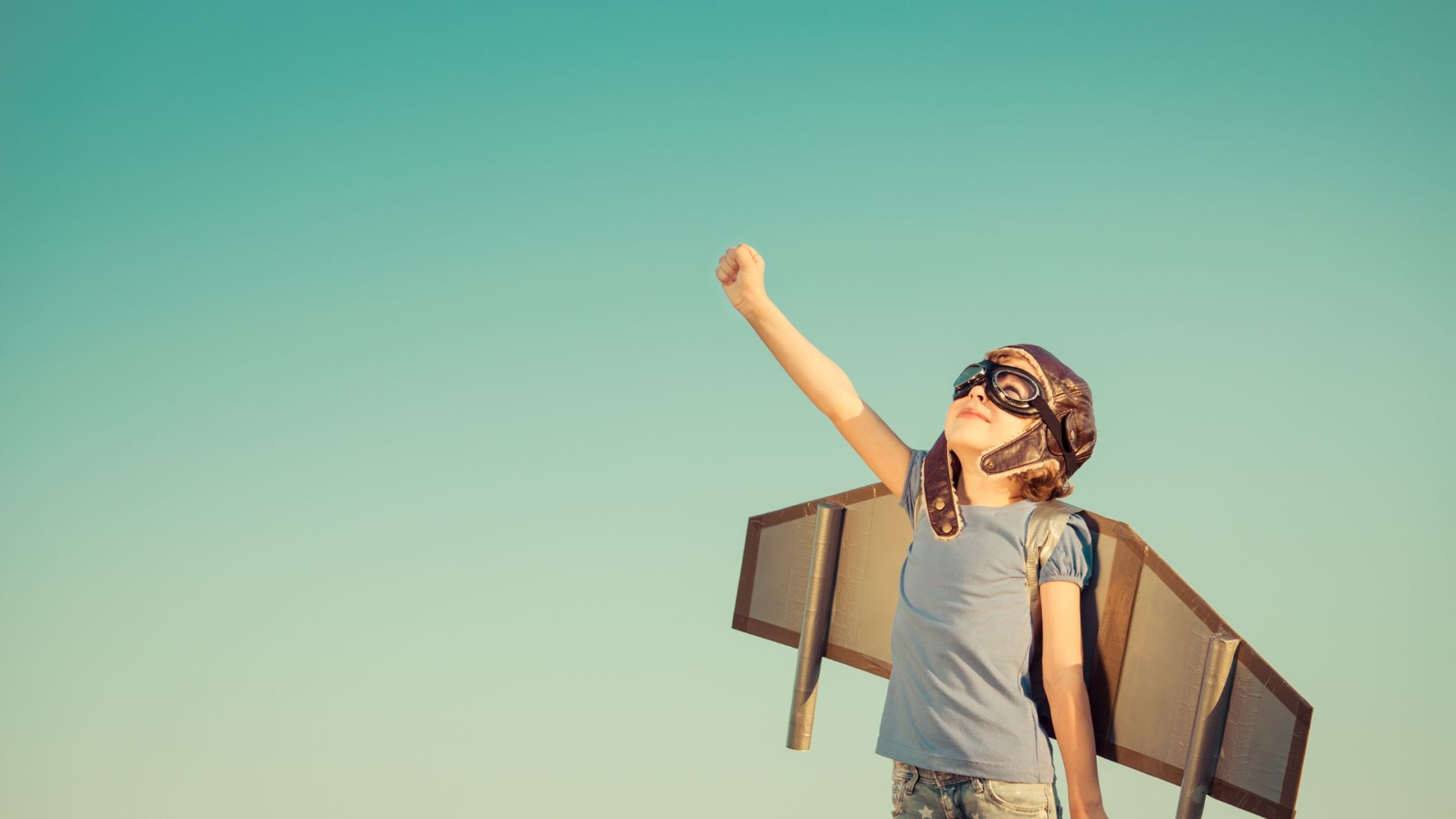 Criança com asas de madeira e capacete de aviação a apontar para o céu