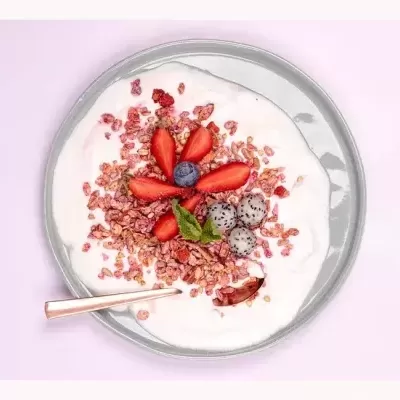 Tigela com iogurte, granola e frutas coloridas, colher dourada e fundo rosa claro