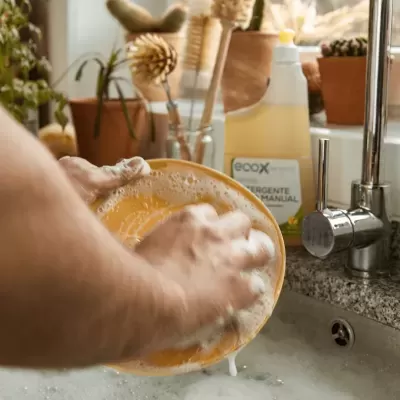 Mãos a lavar prato laranja com detergente ecoX numa pia de granito cinzento com plantas ao fundo