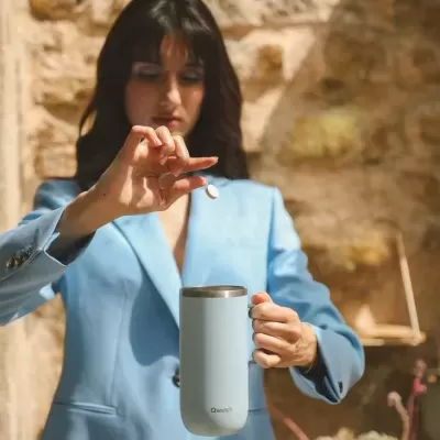 Caneca térmica azul claro com tampa em aço inoxidável sendo segurada por mulher com blazer azul