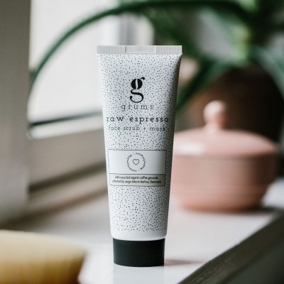 Tubo branco com pontos pretos contendo face scrub + mask 'grums raw espresso' numa superfície branca junto à janela com planta e recipiente rosa ao fundo.