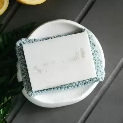 Sabonete branco sobre pano azul e suporte branco com fundo cinzento e folhas verdes