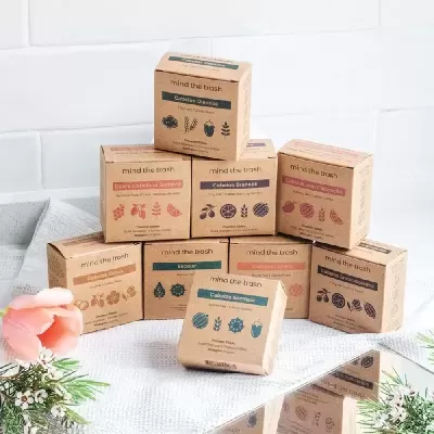 Caixas de sabão sólido mind the trash em embalagem reciclável castanha sobre toalha branca com flores ao lado.