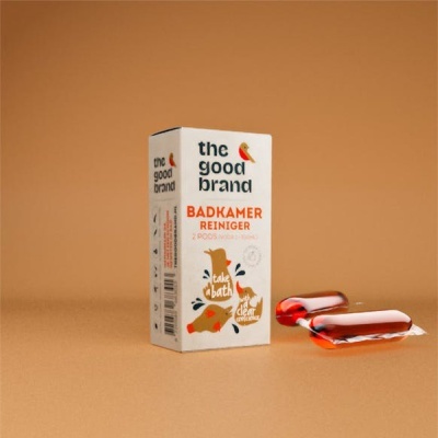 Caixa branca com texto em laranja e preto 'the good brand BADKAMER REINIGER' e dois pods vermelhos em superfície castanha