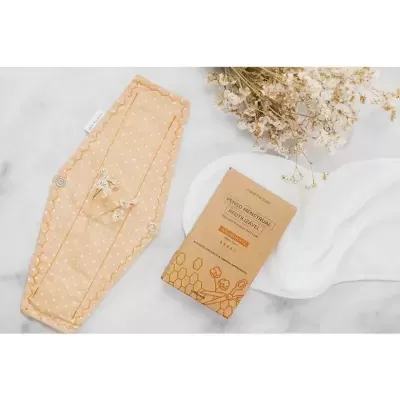 Absorvente reutilizável bege com bolinhas e embalagem castanha, flores secas ao fundo