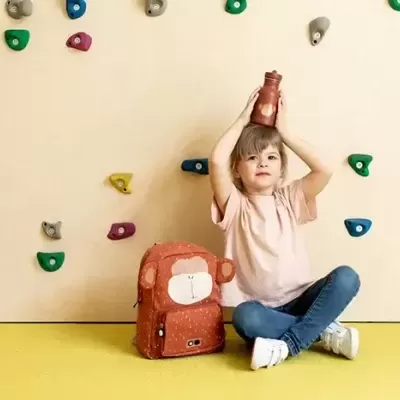 Criança sentada no chão junto à parede de escalada, com mochila laranja e segurando objeto vermelho