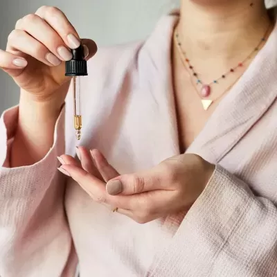 Pessoa usando conta-gotas para aplicar óleo na mão, com roupão vermelho claro e colares.