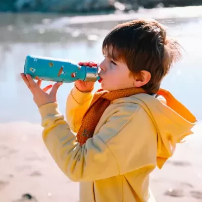 Criança a beber de garrafa térmica azul na praia com casaco amarelo e cachecol castanho