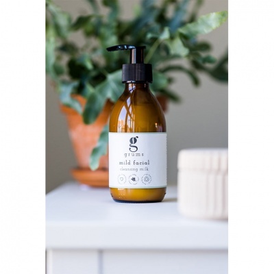 Frasco âmbar com doseador preto e rótulo branco de leite de limpeza facial numa mesa branca com planta ao fundo