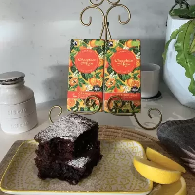Dois pacotes de chocolate com sabor a laranja num suporte dourado, fatia de bolo de chocolate num prato amarelo com limão ao lado.