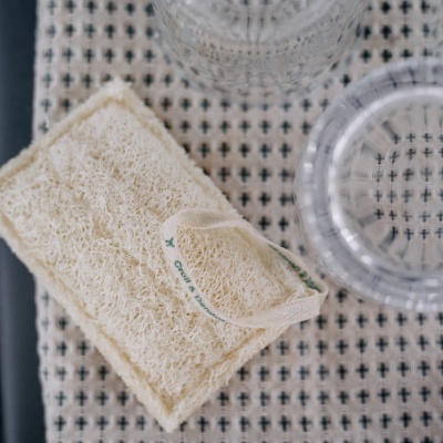 Esponja esfoliante natural com alça branca, pano bege com padrão preto e dois copos de vidro