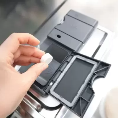 Mão segurando pastilha branca perto de dispensador de detergente cinza