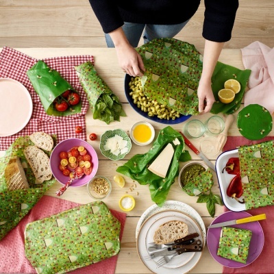 Mesa com alimentos e embalagens reutilizáveis verdes estampadas, prato de massa, pão, queijo e utensílios.
