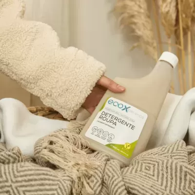 Frasco de detergente líquido para roupa ECOBY sobre mantas bege e castanho com mão vestindo casaco felpudo