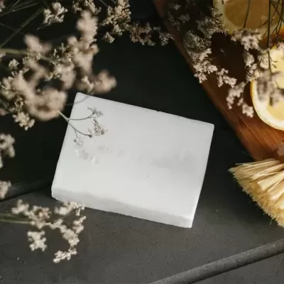 Esponja branca quadrada com pincel de limpeza, flores brancas e fatias de limão sobre superfície cinza e tábua de madeira