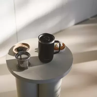 Caneca térmica preta com tampa, infusor de chá metálico e prato com sobremesa sobre mesa redonda cinzenta