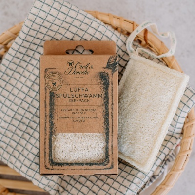 Esponjas de cozinha tipo luffa embaladas em papel cartão dentro de uma cesta de palha, com pano branco de grelha azul por baixo