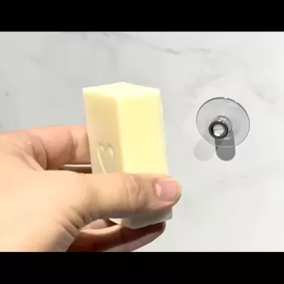 Barra de sabão branca com coração segurada por mão junto a ventosa numa parede branca