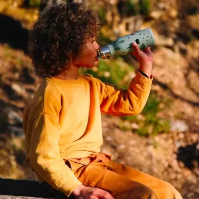 Criança a beber de uma garrafa térmica verde com padrão preto ao ar livre