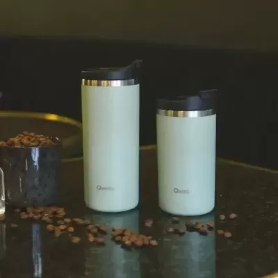 Dois copos térmicos brancos com tampa preta e grãos de café sobre mesa preta.