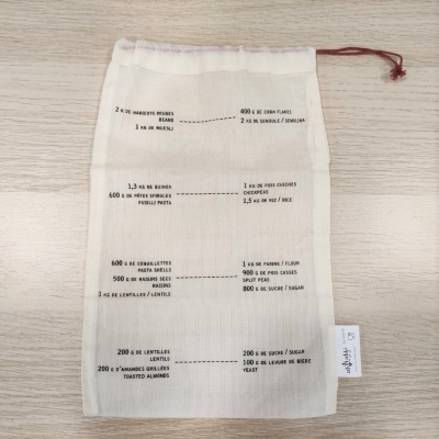 Saco de tecido branco com cordão vermelho e texto impresso em francês sobre uma mesa de madeira clara