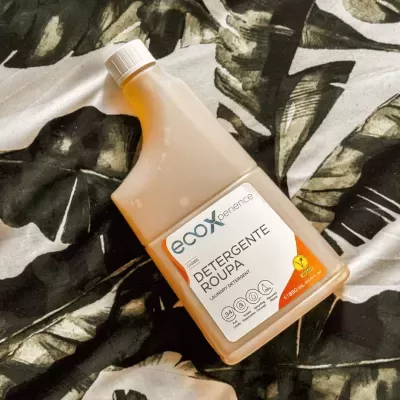 Detergente para roupa ecoXperience numa embalagem branca e laranja sobre tecido com padrão de folhas