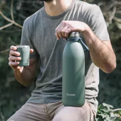 Garrafa térmica verde fosco Qwetch com copo verde nas mãos de uma pessoa vestindo t-shirt cinza.