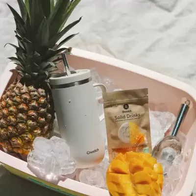 Balde rosa com gelo, abacaxi, copo térmico branco Qwetch, saco Qwetch Solid Drinks, manga cortada.