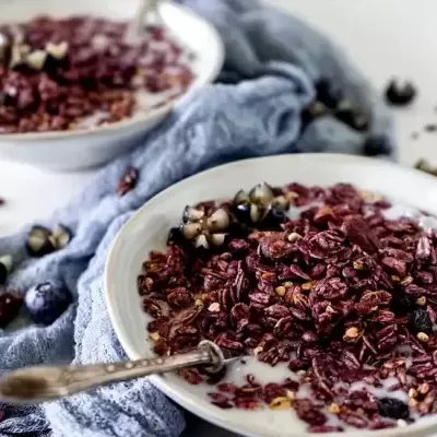 Duas tigelas brancas com granola roxa, frutas secas e líquido branco sobre pano azul