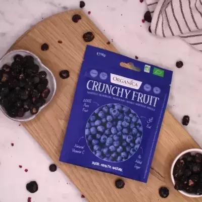 Sacola azul de frutos secos orgânicos sobre tábua de madeira com frutas secas ao redor e fundo de mármore