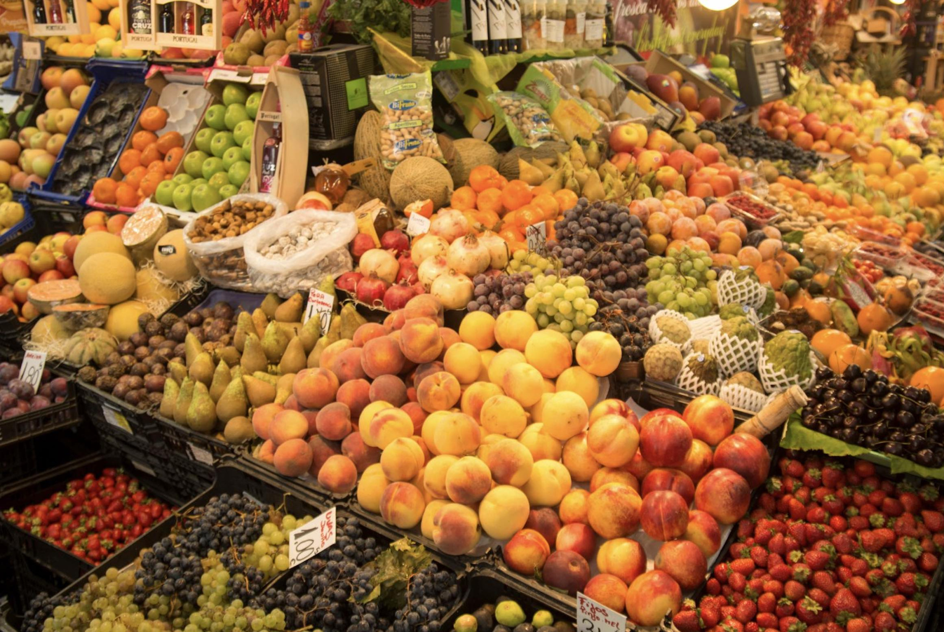 Market stall with assorted fresh fruits and nuts including peaches, grapes, pears, apples, and cantaloupes with visible price tags and wine bottles labeled PORTUGAL.