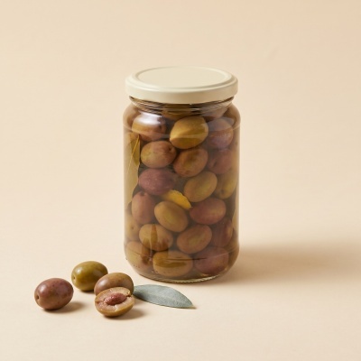 Glass jar filled with mixed olives on beige surface with some loose olives and a leaf