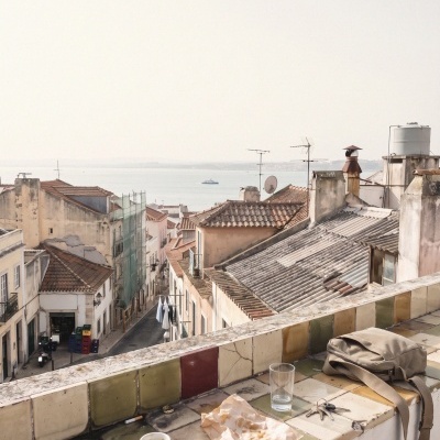Beige shoulder bag on tiled balcony ledge with coffee cup, glass, keys, and paper overlooking rooftops and coastline