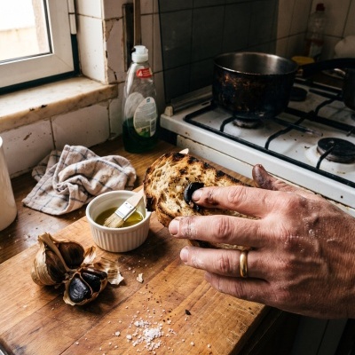 Hand holding toasted bread with black garlic on wooden cutting board in kitchen near stove
