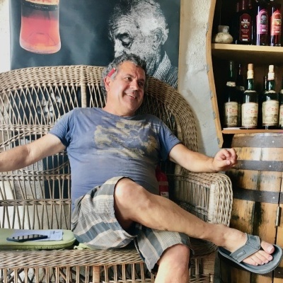 Man sitting on a wicker loveseat with clothing details and beverage bottles on a shelf nearby