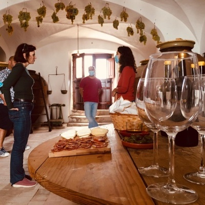 Indoor rustic room with hanging bunches of grapes, people, wine bottles, glasses, and snacks on wooden table