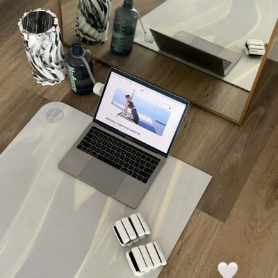 Tapete de yoga branco com padrão cinza, garrafa azul escura, rolo de espuma marmoreado, laptop aberto, pesos brancos e chão de madeira junto a espelho