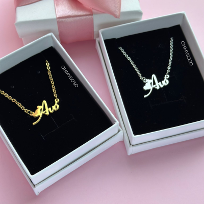 Dois colares com pendente 'Avó' em caixas de joias pretas, uma dourada e uma prateada, caixa branca com fita rosa ao fundo