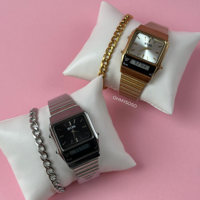 Dois relógios analógico-digitais S135 em metal dourado e prateado com pulseiras tipo corrente, sobre almofadas brancas em fundo rosa.