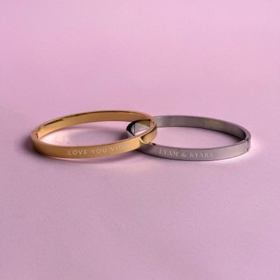 Dois braceletes metálicos, um dourado e um prateado, com gravações, sobre fundo roxo.