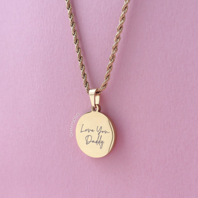 Colar dourado com pendente oval gravado Love You Daddy sobre fundo rosa.