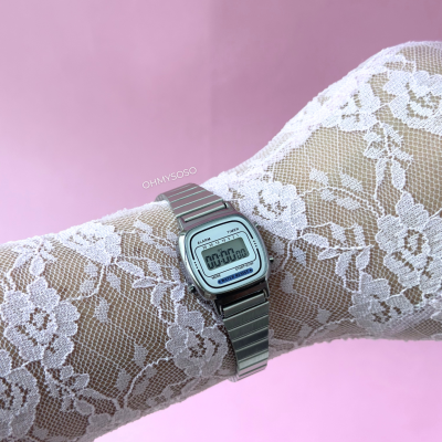 Relógio digital prateado com bracelete metálica sobre tecido rendado branco e fundo rosa