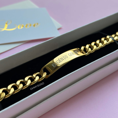 Pulseira dourada com gravação em caixa branca sobre fundo rosa