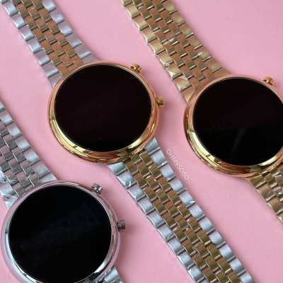 Três relógios de pulso metálicos com mostradores negros em fundo rosa