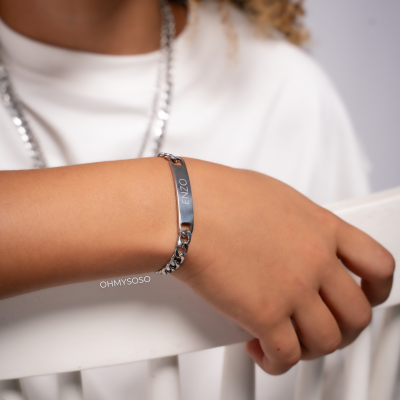 Bracelete prateado com inscrição ENZO usado no pulso de pessoa apoiada numa cadeira branca