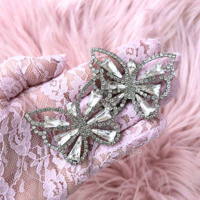 Brincos borboleta prateados com pedras brilhantes na mão com luva de renda rosa sobre fundo felpudo rosa