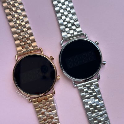 Dois relógios digitais metálicos, um dourado rosado e outro prateado, mostradores pretos, fundo cor rosa.