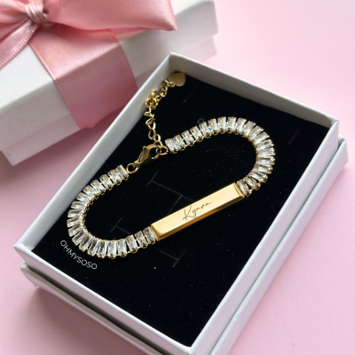 Pulseira dourada com pedras transparentes numa caixa de joias branca com interior preto e caixa de presente branca com fita rosa