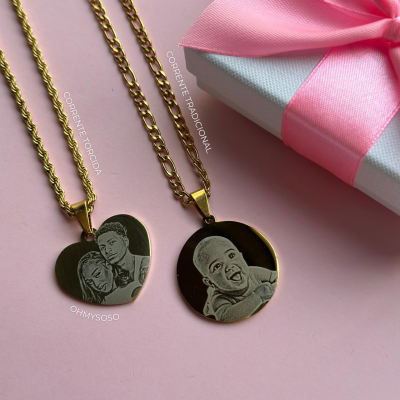 Dois colares dourados com pendentes gravados, um coração e um redondo, sobre fundo rosa com caixa embrulhada em branco e rosa.