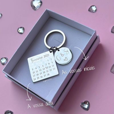Chaveiro metálico com calendário e iniciais numa caixa cinza sobre fundo rosa com pedras em forma de coração